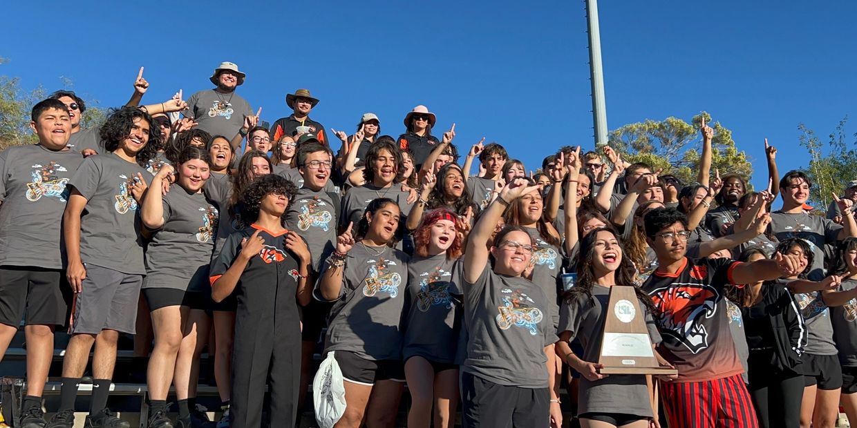 Large group of young people celebrating with a trophy outdoors under clear blue sky.