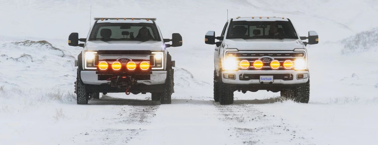 Two white Ford trucks with bright front lights in snowy terrain.