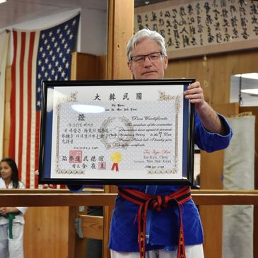 Man in martial arts uniform holding a framed certificate in a dojo.