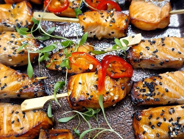 Grilled salmon skewers garnished with black sesame seeds, red chili slices, and fresh herbs.