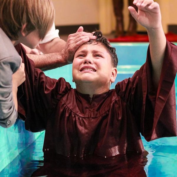 A boy is being baptized in a pool, showing emotion and raised arms.