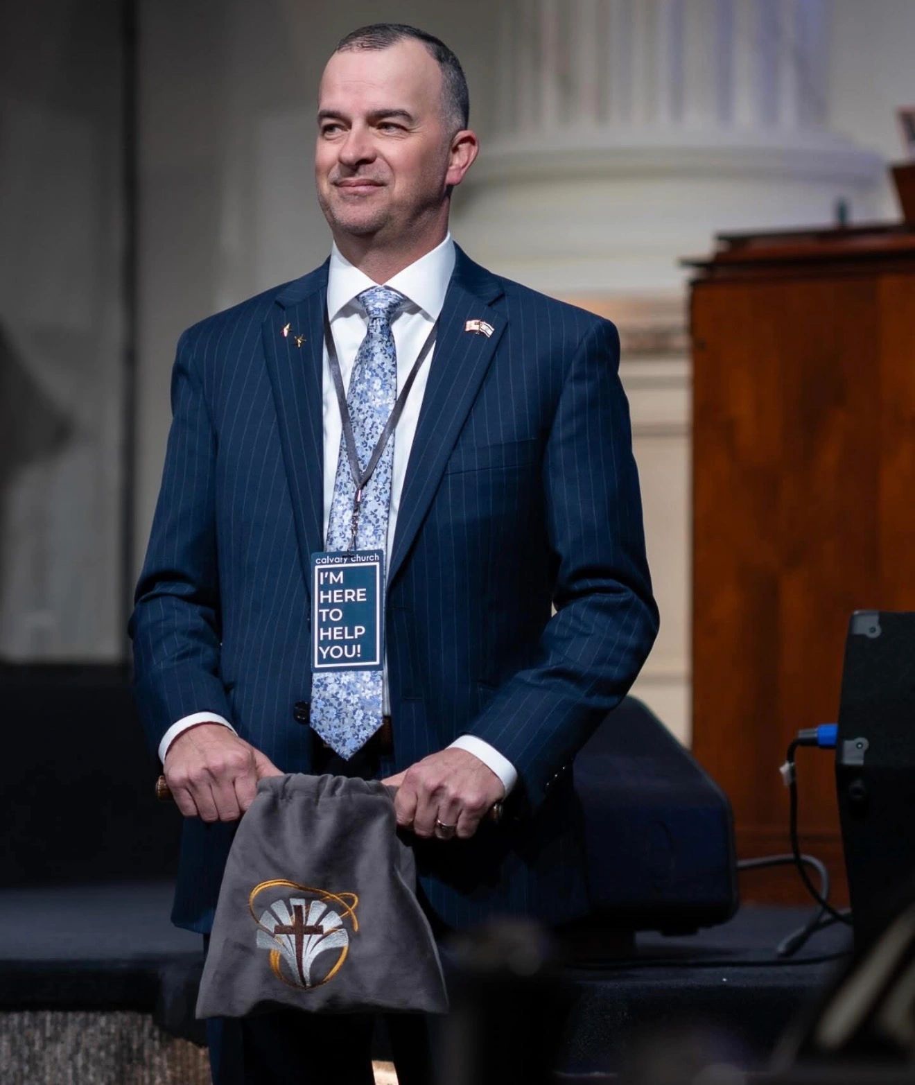 Man in suit with a "I'm here to help you" badge holding a bag with a cross emblem.