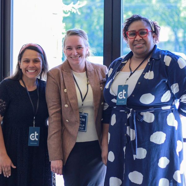 Three smiling women standing together wearing event badges indoors.