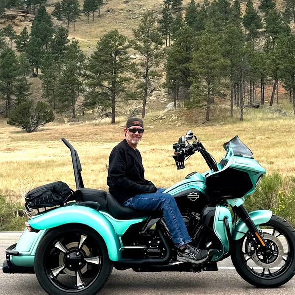 Man sitting on a turquoise Harley-Davidson trike with a rocky butte in the background.