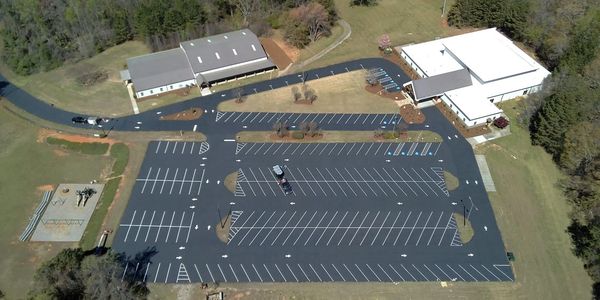 Aerial view of a large, mostly empty parking lot next to two buildings surrounded by trees.