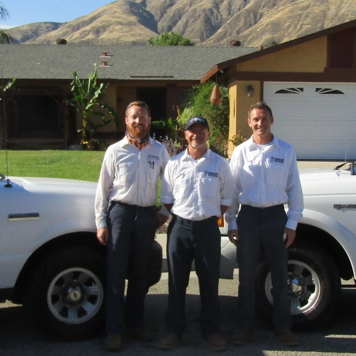 Three men in uniforms standing in front of white trucks and a house.
