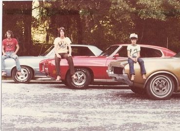 Three boys sit on the hoods of vintage cars in a wooded area.