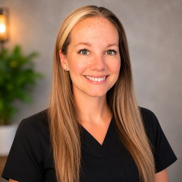 Smiling woman with long blonde hair in black scrubs against a neutral background.