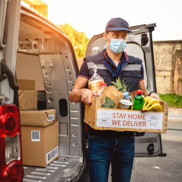 Delivery man with groceries wearing a face mask and holding a crate saying 'Stay Home We Deliver'.