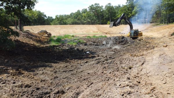 Excavator digging in a muddy construction site surrounded by trees.