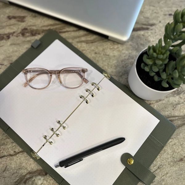 Open notebook with glasses and pen on a stone desk beside a laptop and plant.