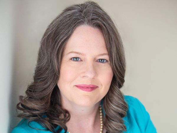 Confident woman in a turquoise jacket with folded arms and a pearl necklace.