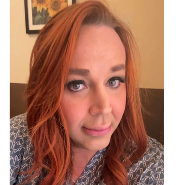 Woman with red hair and soft makeup smiling gently indoors.