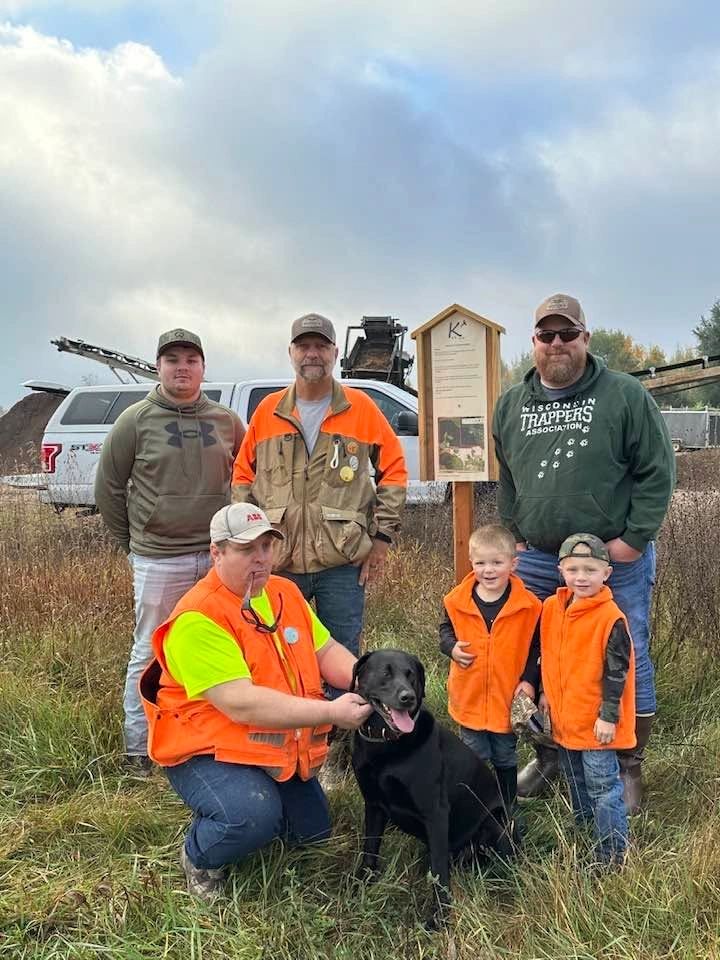 Group of men, two kids in orange vests, and a black dog outdoors.
