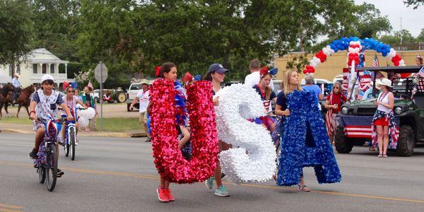 Three people walk in a parade carrying large letters that spell out USA.