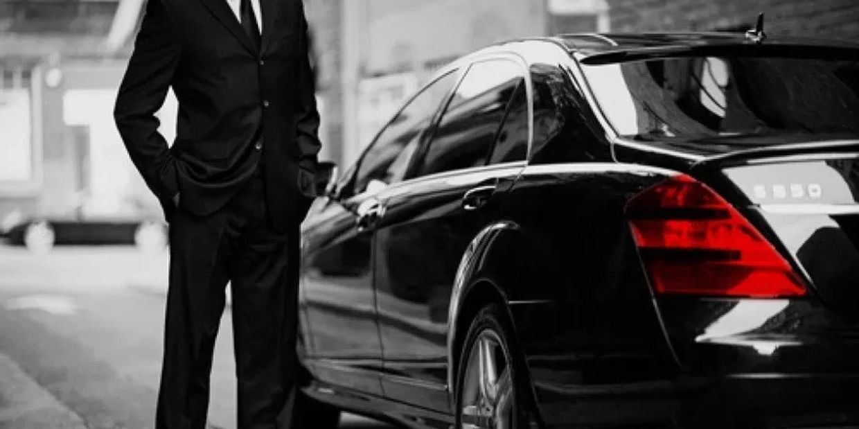 Man in black suit standing next to a luxury black car.