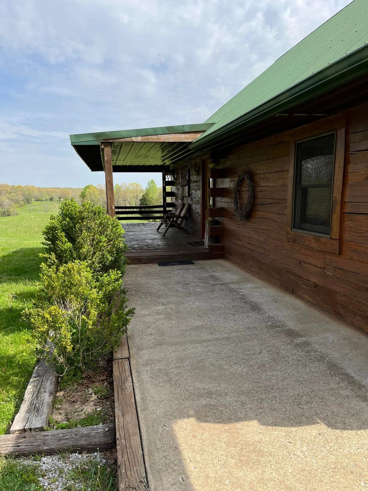 A wooden cabin with a green roof and porch, surrounded by bushes and a grassy field.