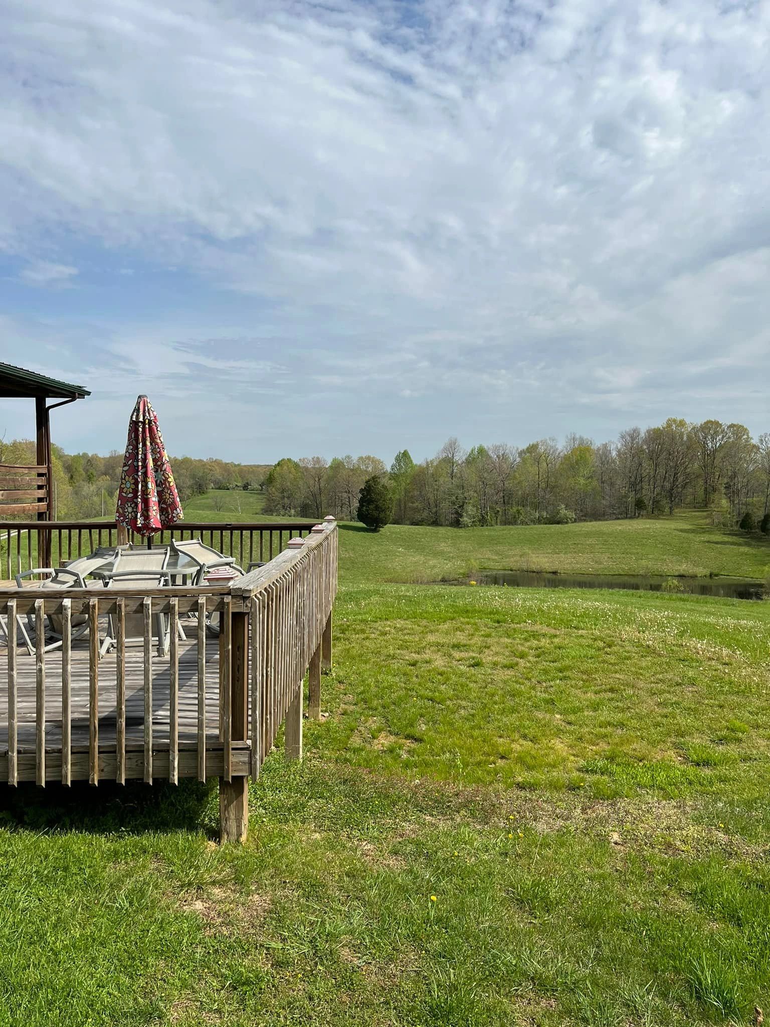 A wooden deck with furniture and an umbrella overlooks a grassy field with a pond and trees in the background.