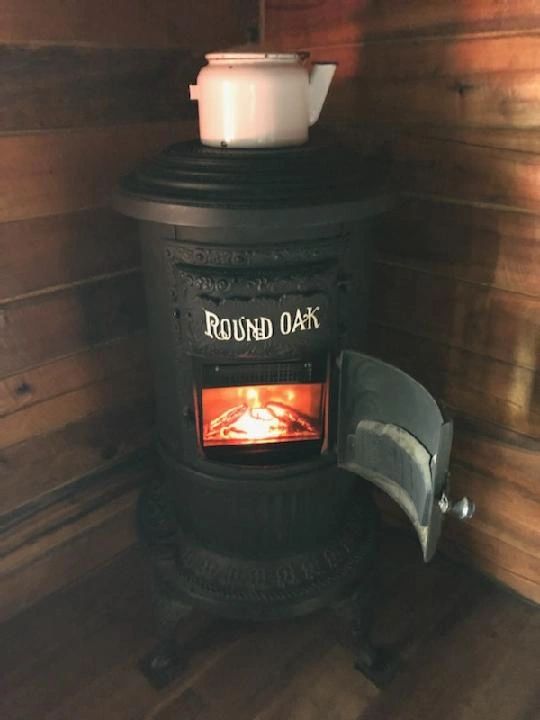 A lit wood-burning stove with a tea kettle on top, in a wooden room.