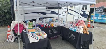 Outdoor Scentsy product display with candles and plush toys under a canopy.