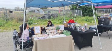 Two women sitting under tents at an outdoor market stall with various items for sale.