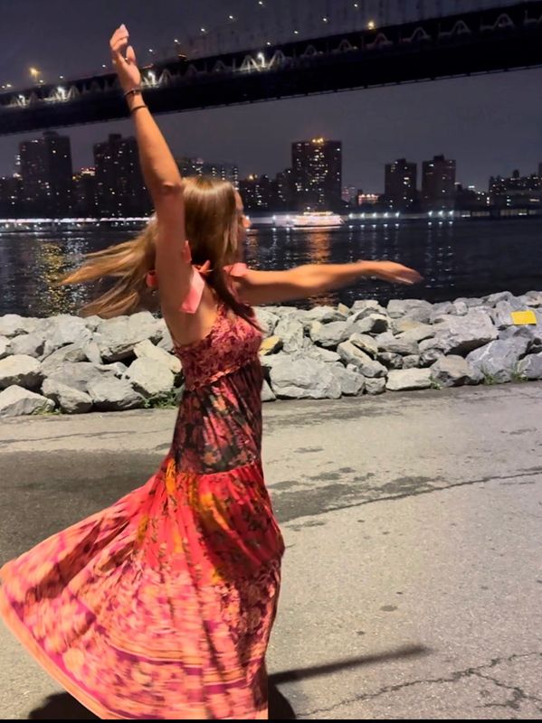 Woman in a vibrant dress twirling by the waterfront at night under a lit bridge.