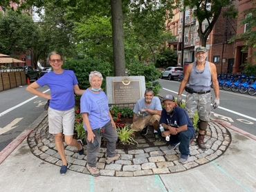 Five people posing by a tree with a historical plaque in an urban area.