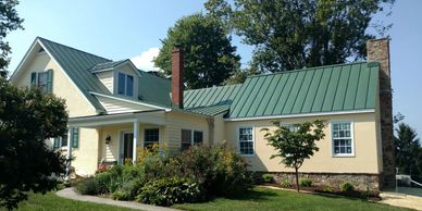 A charming house with a green metal roof and cream-colored walls surrounded by greenery.
