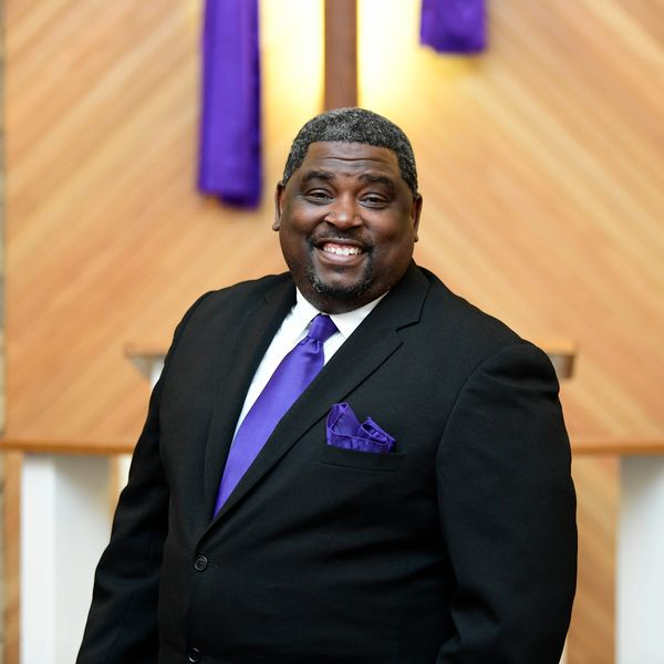 A smiling man in a black suit with purple accents stands indoors.