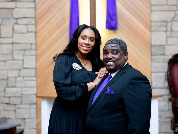 A smiling couple dressed formally in front of a wooden cross with purple cloth.