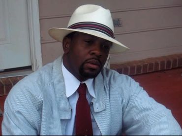 Man wearing a white hat, red tie, and striped jacket sitting outdoors.
