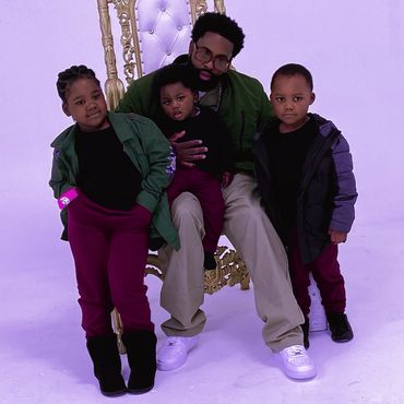 A man with three children poses in front of a decorative chair in a studio.