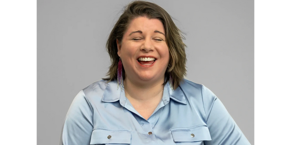 Smiling woman in light blue shirt with colorful earrings against gray background.
