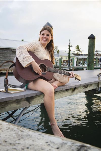 Young woman playing guitar and smiling on a dock by the water.