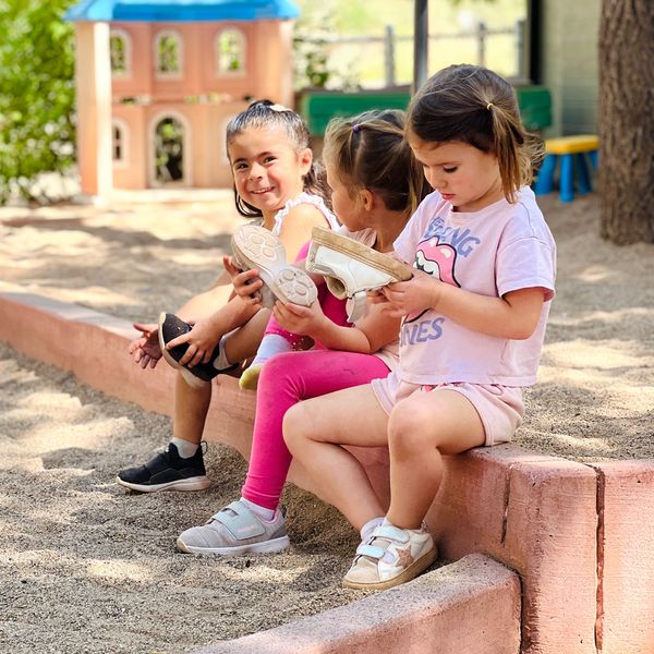 Three young girls sitting by a sandbox, holding their shoes and enjoying the outdoors.