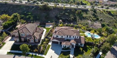 Aerial view of two large houses with pools and greenery in a suburban area.