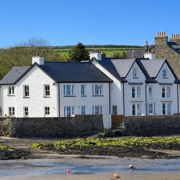 White coastal houses behind a stone wall with a green hill and blue sky.