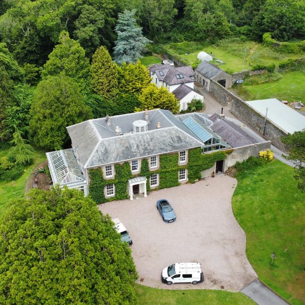 A large ivy-covered country house surrounded by greenery with cars parked outside.