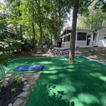 Backyard with green artificial turf surrounded by trees and a tree in the middle.
