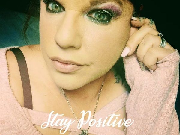 Close-up of a woman with green eyes and makeup, wearing a pink sweater and heart pendant necklace.