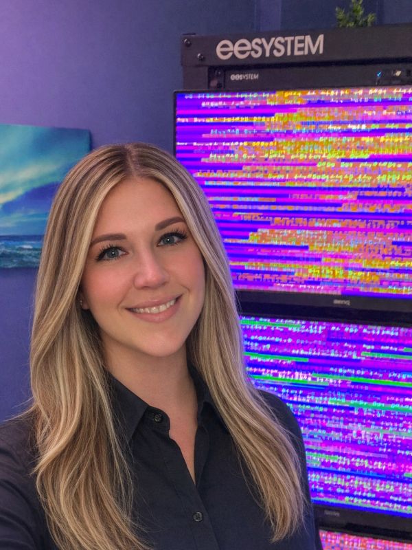Smiling woman with long blonde hair in front of glitchy monitors.