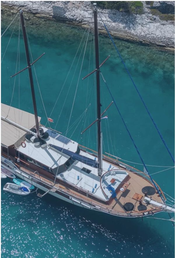 A large sailboat anchored near a rocky shore in clear blue water.
