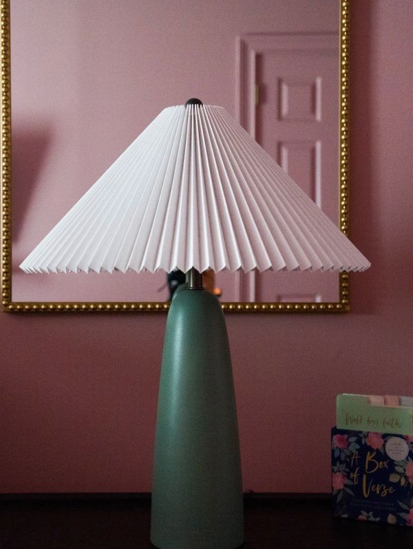 A green lamp with a pleated white lampshade against a pink wall.