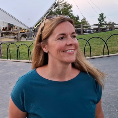 A woman smiling outdoors near a park with a large white structure in the background.