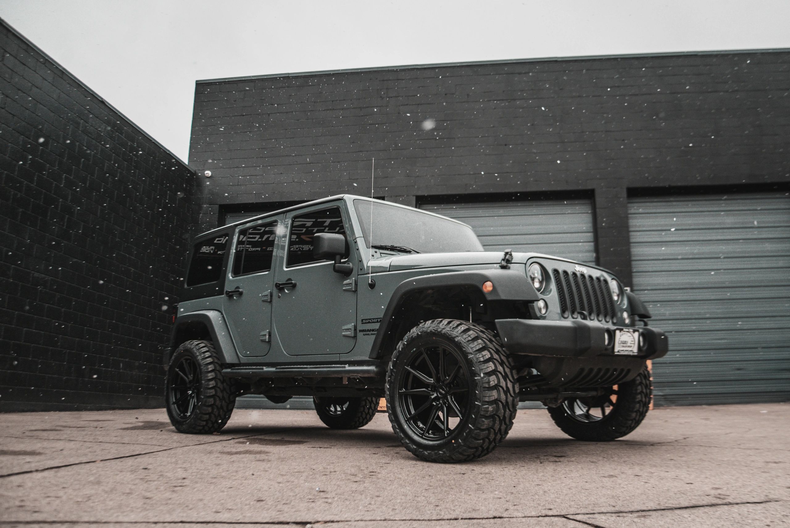 Matte gray Jeep Wrangler parked outside a black building during snowfall.