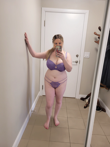 Woman taking a mirror selfie wearing purple lingerie in a small room.