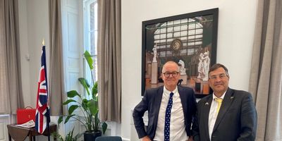 Two men in suits standing in a formal room with a British flag and plants.