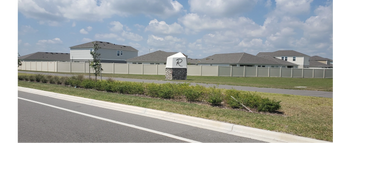 A suburban neighborhood entrance with a stone sign and houses behind a fence.