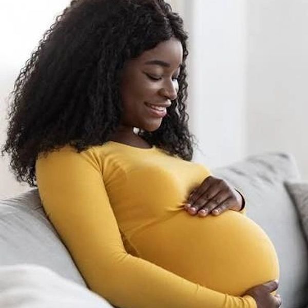 Smiling pregnant woman in a yellow top sitting on a couch, cradling her belly.