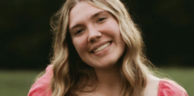 Young woman with blonde hair smiling outdoors in a pink top.